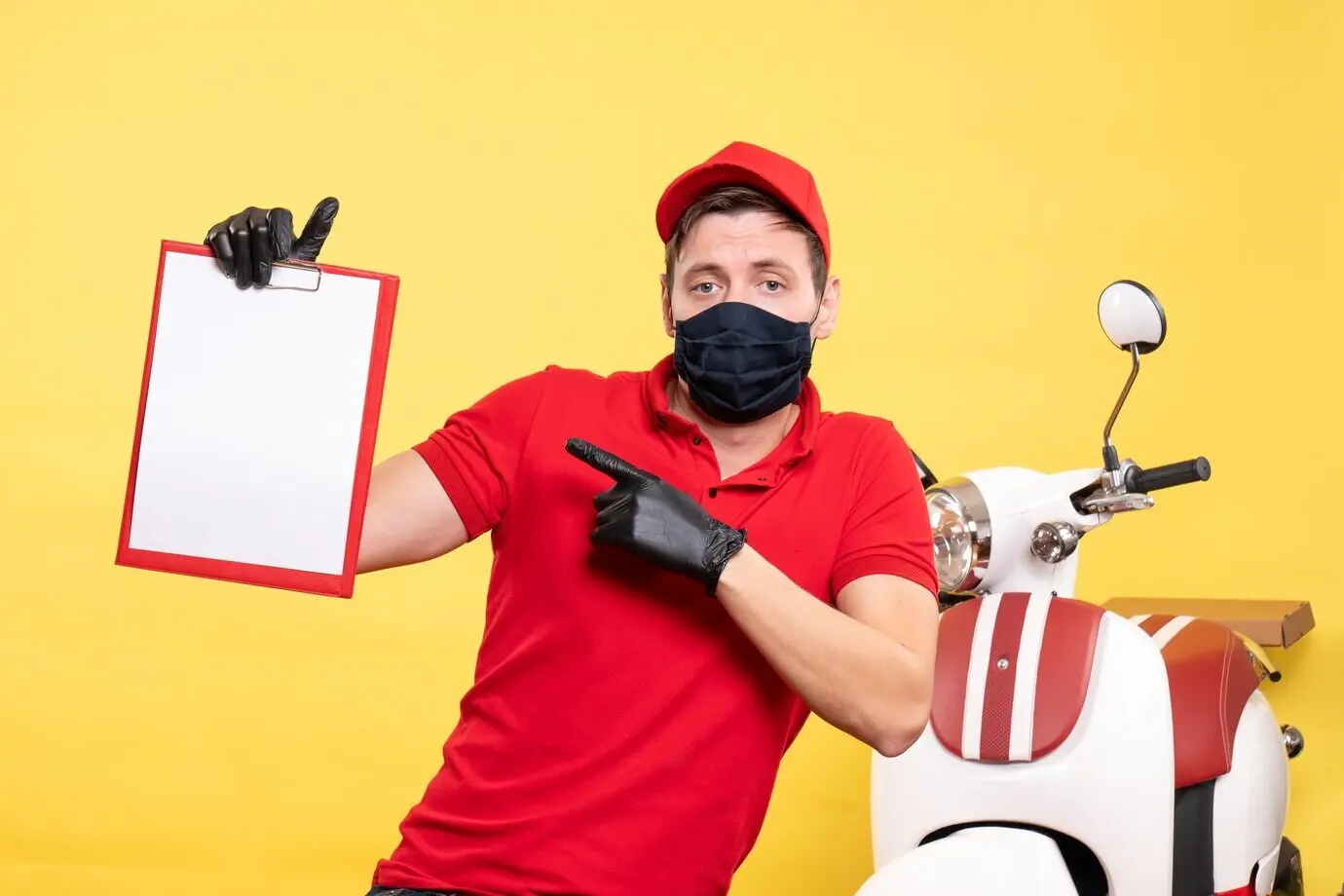 Фронтальный вид: курьер-мужчина в чёрной маске с папкой, в жёлтой униформе; работа сервиса доставки во время пандемии COVID.