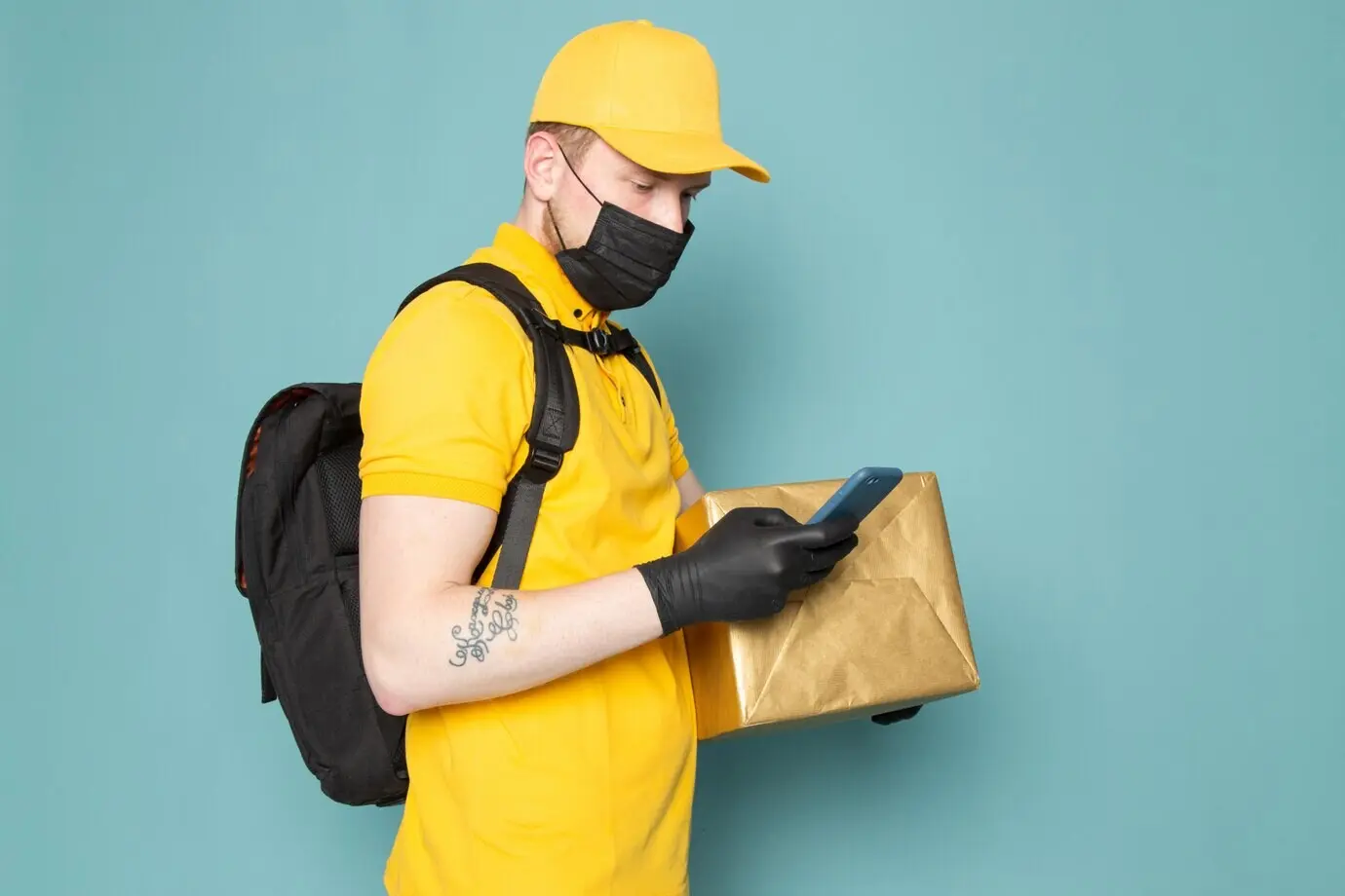 Молодой курьер в желтом поло, желтой кепке, белых джинсах, с рюкзаком и в черной стерильной маске держит коробку на синем фоне.